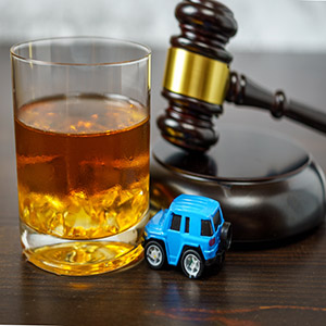 A glass of whiskey next to a judge's gavel and a small blue toy car: legal consequences of a scooter DUI.